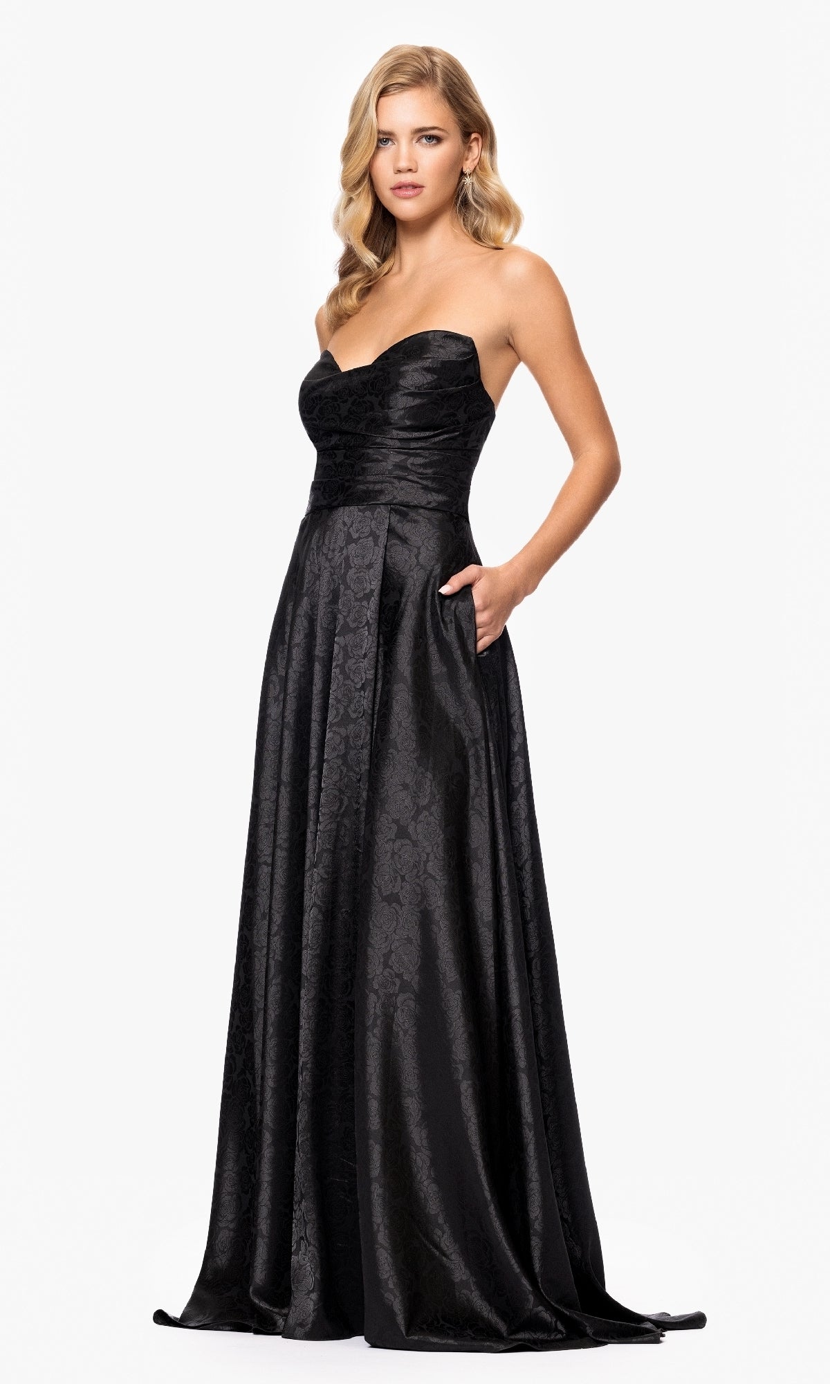 A woman models the Strapless Black Prom Gown: Blondie Nites 5301BN, featuring a subtle floral pattern, against a plain white background.